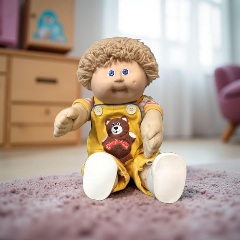K&ouml;nnte beinhalten: Eine Cabbage Patch Kids-Puppe mit blondem, lockigem Haar und blauen Augen. Die Puppe tr&auml;gt einen gelben Overall mit einem braunen B&auml;renaufn&auml;her und wei&szlig;en Schuhen. Die Puppe sitzt auf einem rosa Teppich.