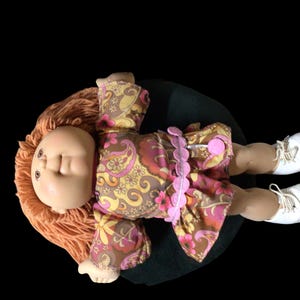 Puede incluir: Una mu&ntilde;eca vintage con cabello naranja y una cara pintada, vestida con un vestido estampado floral en tonos marr&oacute;n, rosa y amarillo. La mu&ntilde;eca tiene zapatos blancos y est&aacute; acostada sobre una superficie verde oscuro.