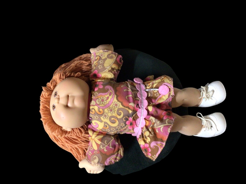 Puede incluir: Una mu&ntilde;eca vintage con cabello naranja y una cara pintada, vestida con un vestido estampado floral en tonos marr&oacute;n, rosa y amarillo. La mu&ntilde;eca tiene zapatos blancos y est&aacute; acostada sobre una superficie verde oscuro.