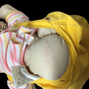 K&ouml;nnte beinhalten: Eine Vintage-Puppe mit blonden Garnhaaren, die eine gelbe Jacke und ein gestreiftes Hemd in Rosa, Gelb und Wei&szlig; tr&auml;gt. Der K&ouml;rper der Puppe besteht aus einem hellbeigen Stoff. Ein Etikett ist sichtbar.