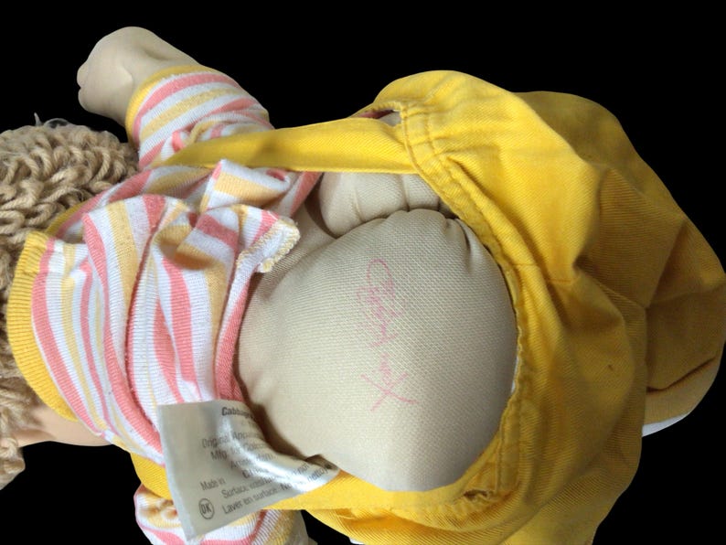 K&ouml;nnte beinhalten: Eine Vintage-Puppe mit blonden Garnhaaren, die eine gelbe Jacke und ein gestreiftes Hemd in Rosa, Gelb und Wei&szlig; tr&auml;gt. Der K&ouml;rper der Puppe besteht aus einem hellbeigen Stoff. Ein Etikett ist sichtbar.
