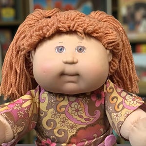 Puede incluir: Una mu&ntilde;eca Cabbage Patch Kid vintage con cabello de hilo color melocot&oacute;n y ojos azules. La mu&ntilde;eca viste un vestido con estampado floral en tonos marr&oacute;n, rosa y amarillo. Los brazos y la cara de la mu&ntilde;eca son de material suave.