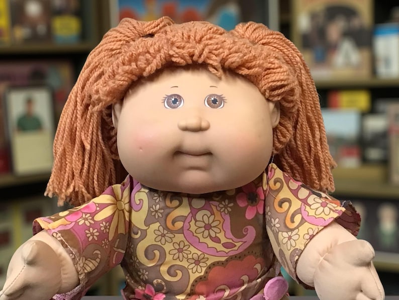 Puede incluir: Una mu&ntilde;eca Cabbage Patch Kid vintage con cabello de hilo color melocot&oacute;n y ojos azules. La mu&ntilde;eca viste un vestido con estampado floral en tonos marr&oacute;n, rosa y amarillo. Los brazos y la cara de la mu&ntilde;eca son de material suave.