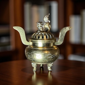 May include: A vintage brass incense burner with a decorative lion-like figure on top. The burner has a round body with etched cloud designs and three ornate feet. It features curved handles and a perforated lid.