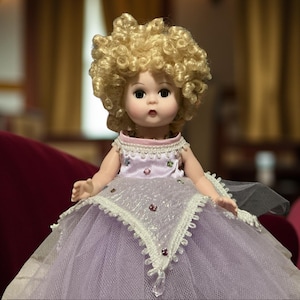 Peut inclure: Poupée vintage aux cheveux blonds bouclés et au visage délicat. La poupée porte une robe lavande avec une jupe en tulle, ornée de dentelle blanche et de bijoux étincelants. La poupée est assise sur une surface en velours bordeaux.