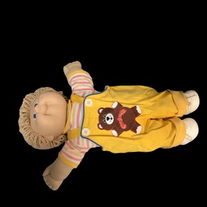 K&ouml;nnte beinhalten: Vintage Cabbage Patch Kid Puppe, liegend auf schwarzem Hintergrund. Die Puppe hat blonde Garnhaare, blaue Augen und tr&auml;gt einen gelben Overall mit einem braunen B&auml;ren-Applikation. Die &Auml;rmel sind gestreift.