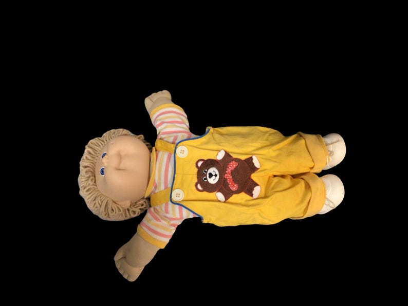 K&ouml;nnte beinhalten: Vintage Cabbage Patch Kid Puppe, liegend auf schwarzem Hintergrund. Die Puppe hat blonde Garnhaare, blaue Augen und tr&auml;gt einen gelben Overall mit einem braunen B&auml;ren-Applikation. Die &Auml;rmel sind gestreift.