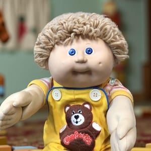 K&ouml;nnte beinhalten: Vintage Cabbage Patch Kid Puppe mit blauen Augen und blondem Garnhaar. Die Puppe tr&auml;gt einen gelben Overall mit einem braunen B&auml;renaufn&auml;her und ein gestreiftes Hemd darunter. Ein klassisches Spielzeug.