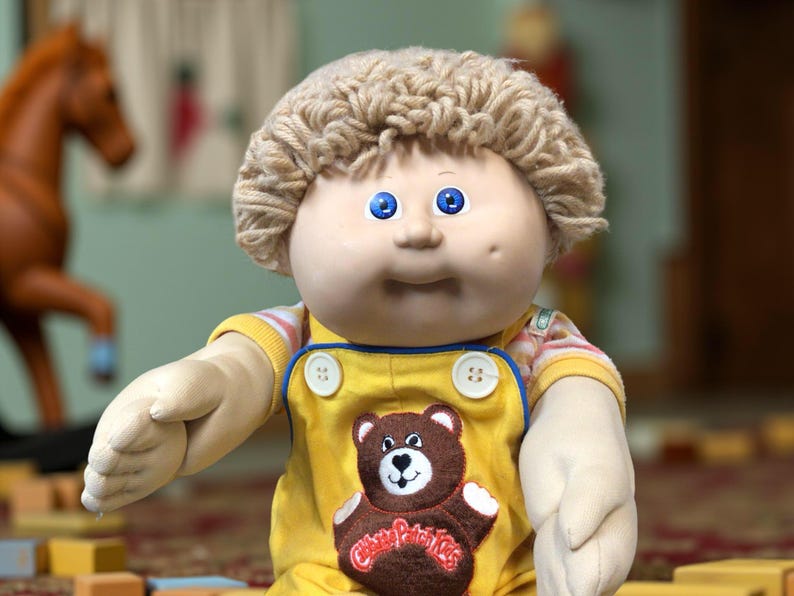 K&ouml;nnte beinhalten: Vintage Cabbage Patch Kid Puppe mit blauen Augen und blondem Garnhaar. Die Puppe tr&auml;gt einen gelben Overall mit einem braunen B&auml;renaufn&auml;her und ein gestreiftes Hemd darunter. Ein klassisches Spielzeug.