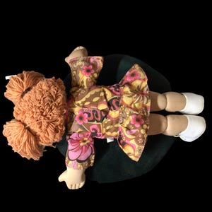 Puede incluir: Mu&ntilde;eca hecha a mano con pelo de hilo naranja y un vestido con estampado floral en tonos marrones, amarillos y rosas. La mu&ntilde;eca tiene zapatos blancos y est&aacute; tumbada sobre una superficie verde oscuro.