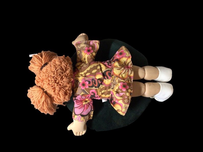 Puede incluir: Mu&ntilde;eca hecha a mano con pelo de hilo naranja y un vestido con estampado floral en tonos marrones, amarillos y rosas. La mu&ntilde;eca tiene zapatos blancos y est&aacute; tumbada sobre una superficie verde oscuro.