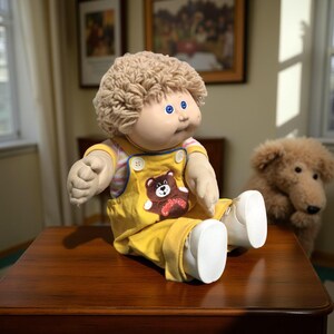 K&ouml;nnte beinhalten: Eine Vintage Cabbage Patch Kid-Puppe mit blondem, lockigem Haar, blauen Augen und einem runden Gesicht. Sie tr&auml;gt einen gelben Overall mit einem B&auml;ren-Applikation und wei&szlig;en Schuhen. Ein Stoffhund sitzt im Hintergrund.