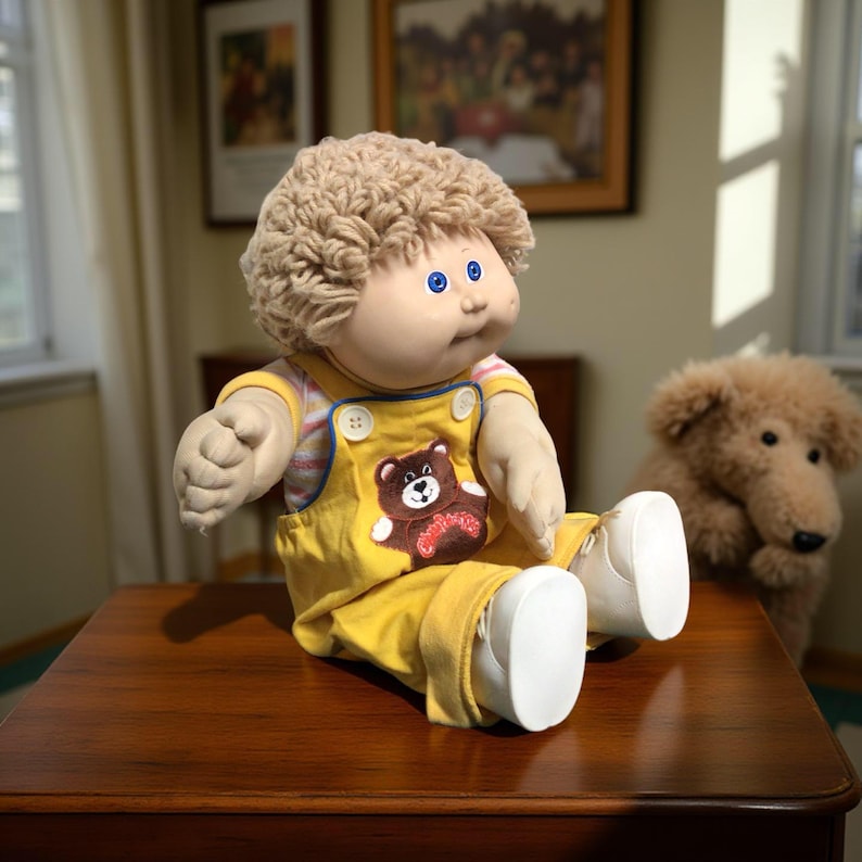 K&ouml;nnte beinhalten: Eine Vintage Cabbage Patch Kid-Puppe mit blondem, lockigem Haar, blauen Augen und einem runden Gesicht. Sie tr&auml;gt einen gelben Overall mit einem B&auml;ren-Applikation und wei&szlig;en Schuhen. Ein Stoffhund sitzt im Hintergrund.
