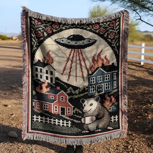 May include: Woven throw blanket with a black border and fringe. The design features a UFO, burning houses, and an opossum holding a mug. The top border has a floral pattern. The background is a light beige color.