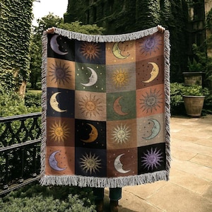 May include: A woven tapestry blanket featuring a celestial design with sun and moon motifs in various colors. The blanket has a fringed edge and is displayed outdoors. The design includes squares with suns and crescent moons.