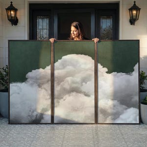 May include: A triptych of framed art panels depicting a cloudscape. The artwork has a dark green background with a large, fluffy white cloud formation. The frames are dark brown. The panels are being held by a person.