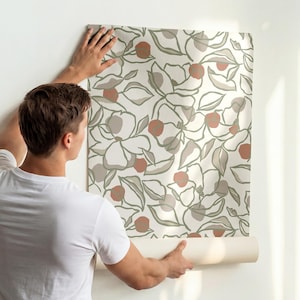 Könnte beinhalten: Eine Tapetenrolle mit einem sich wiederholenden botanischen Muster aus umrandeten Blumen und Blättern in Salbeigrün, Creme und Terrakotta-Orange. Die Tapete wird auf eine schlichte weiße Wand über einer salbeigrünen Wandverkleidung angebracht.