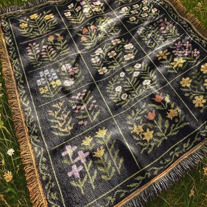 May include: A black woven blanket with a floral pattern in shades of pink, yellow, and white. The blanket has a grid design and a fringed edge. The blanket is placed on a grassy surface.