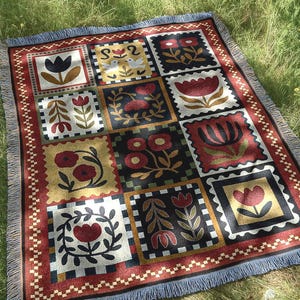 May include: A rectangular throw blanket with a floral pattern in a grid design. The blanket features squares with various flower illustrations in red, black, and yellow, framed by a red border and blue fringe. The blanket is placed on a grassy surface.