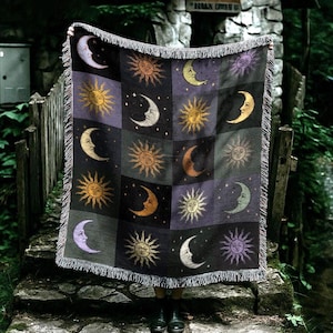 May include: A woven throw blanket featuring a celestial design of moons and suns in shades of gold, purple, and black. The blanket has a fringed edge and is held up in front of a stone structure and wooden fence.