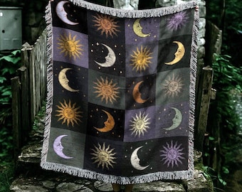 Peut inclure: Une couverture tissée avec un motif céleste de lunes et de soleils en or, violet et noir. La couverture a une bordure à franges. Elle est tenue devant une structure en pierre et une clôture en bois.