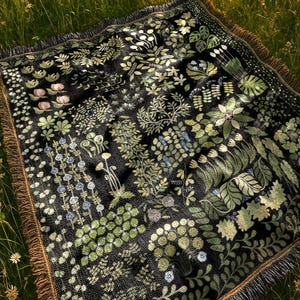 May include: A black woven blanket featuring a botanical design of green and light green foliage and flowers. The blanket has a brown fringed edge and is shown on a grassy surface.