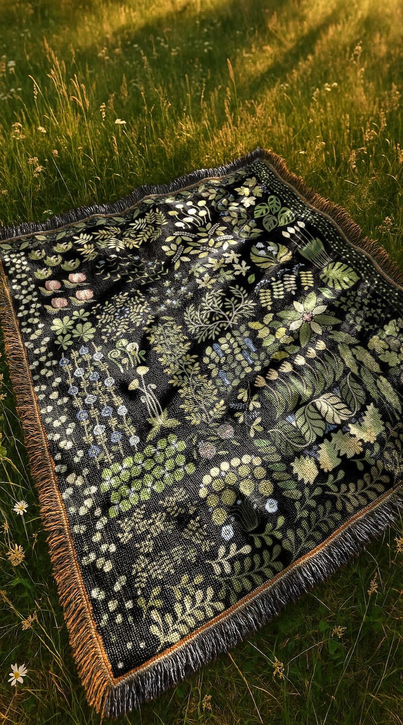May include: A black woven blanket featuring a botanical design of green and light green foliage and flowers. The blanket has a brown fringed edge and is shown on a grassy surface.