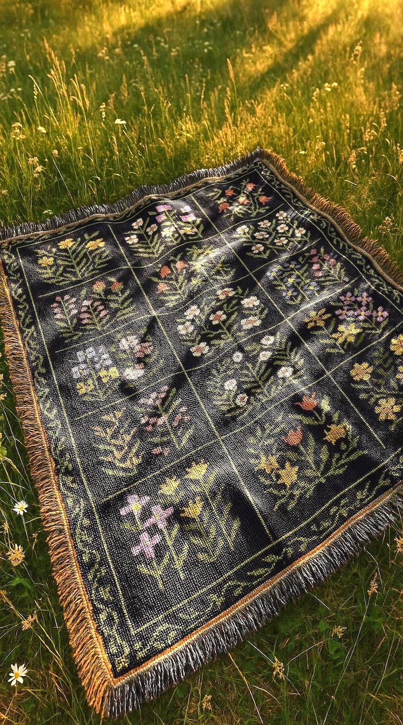 May include: A black woven blanket with a floral pattern in shades of pink, yellow, and white. The blanket has a grid design and a fringed edge. The blanket is placed on a grassy surface.