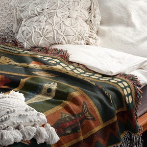 Puede incluir: Una manta tejida con un dise&ntilde;o de pez en tonos verdes, marrones y dorados, bordes con flecos y una almohada de macram&eacute;. La manta est&aacute; sobre una cama con ropa de cama blanca y una almohada decorativa.