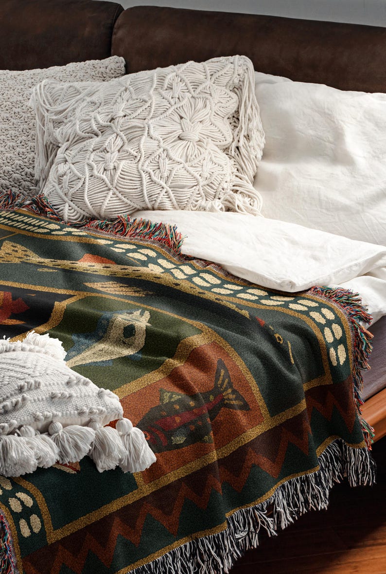 Puede incluir: Una manta tejida con un dise&ntilde;o de pez en tonos verdes, marrones y dorados, bordes con flecos y una almohada de macram&eacute;. La manta est&aacute; sobre una cama con ropa de cama blanca y una almohada decorativa.