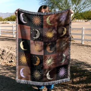 Peut inclure: Une couverture tissée avec un motif répété de soleils et de croissants de lune. Les carrés alternent entre des fonds noirs, bruns et beiges. La couverture a une bordure à franges. La couverture est tenue devant une personne.