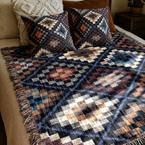 May include: A woven blanket and two matching throw pillows with a geometric pattern in shades of black, brown, blue, and red. The blanket has a fringed edge. The pillows are square and placed on a bed.