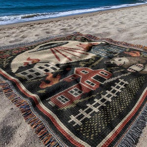 May include: A woven blanket with a rural scene design, featuring a red barn, houses, and a fence. The blanket has a black background with a cream-colored sky and is fringed with orange, blue, and black threads. It is placed on a sandy beach.