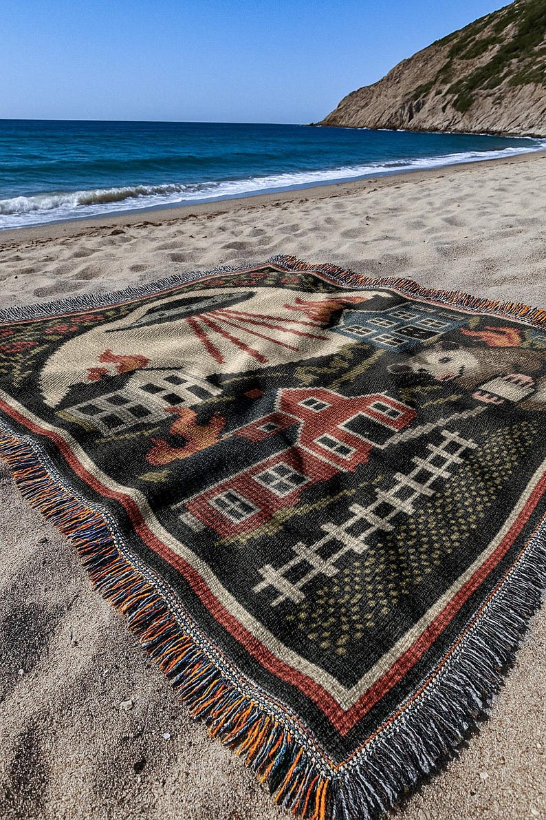 May include: A woven blanket with a rural scene design, featuring a red barn, houses, and a fence. The blanket has a black background with a cream-colored sky and is fringed with orange, blue, and black threads. It is placed on a sandy beach.