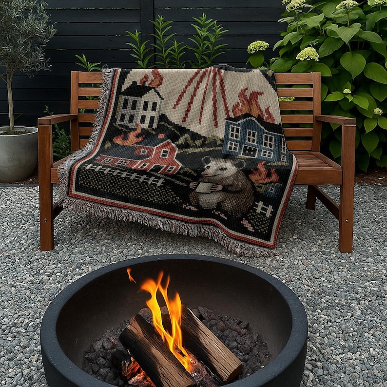 May include: A woven throw blanket with a whimsical design featuring houses on fire, a possum, and a rural landscape. The blanket is draped over a wooden bench. The color palette includes black, red, beige, and brown.