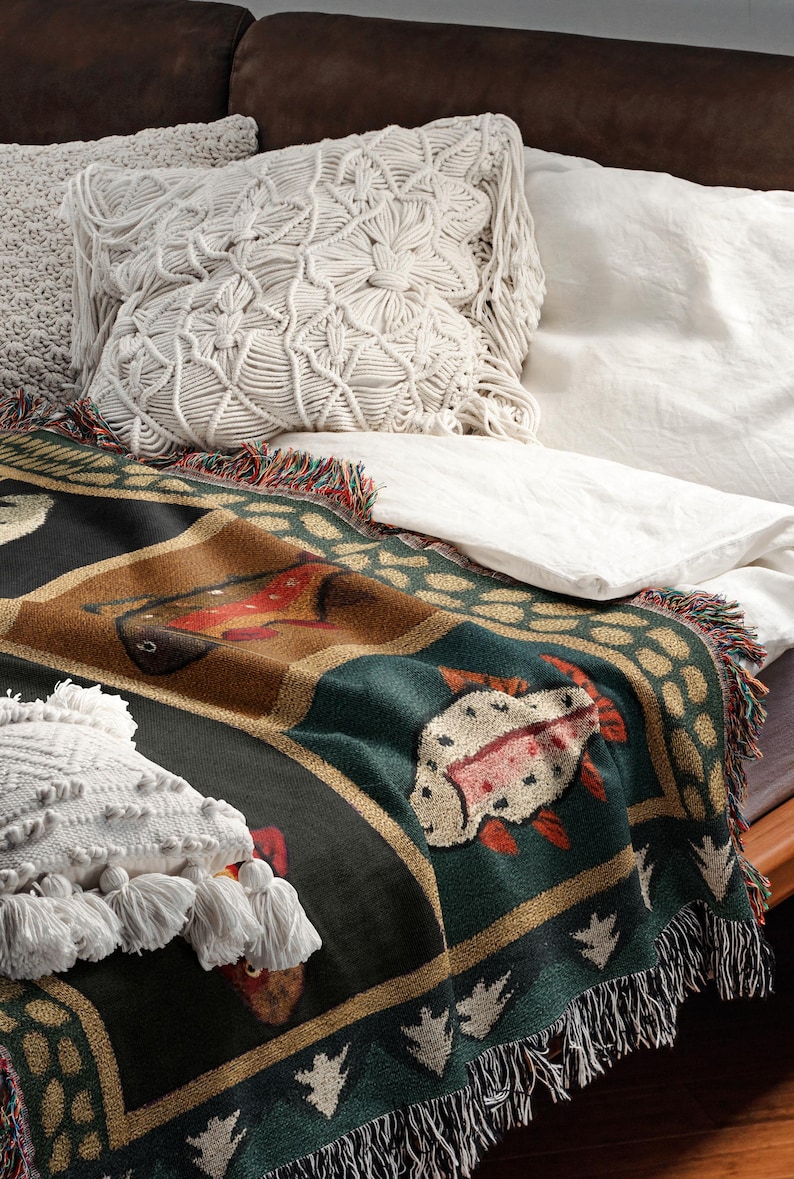 Puede incluir: Una manta tejida con un dise&ntilde;o de pez en tonos verdes, marrones y rojos. La manta tiene un borde con flecos y est&aacute; colocada sobre una cama con s&aacute;banas blancas y cojines decorativos.