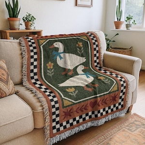 May include: A woven throw blanket featuring two white geese with blue bows on a green background. The blanket has a decorative border with a checkered pattern and fringe. The blanket is draped over a beige sofa.