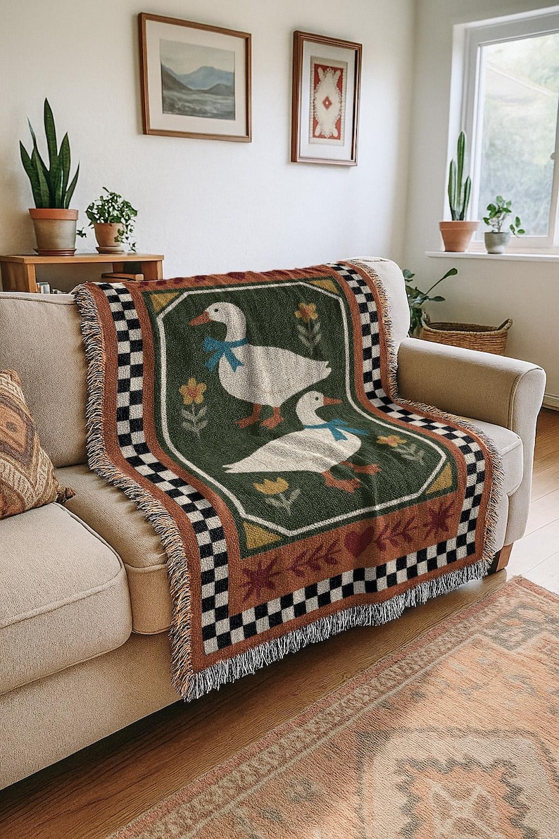May include: A woven throw blanket featuring two white geese with blue bows on a green background. The blanket has a decorative border with a checkered pattern and fringe. The blanket is draped over a beige sofa.