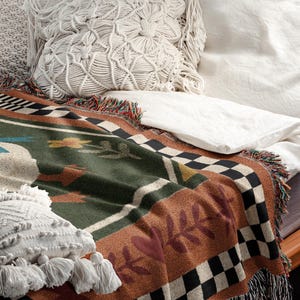 May include: A bed with a patterned throw blanket and decorative pillows. The blanket features a green, brown, and black design with a checkered border and fringe. Two white macrame pillows and a white sheet are also visible.