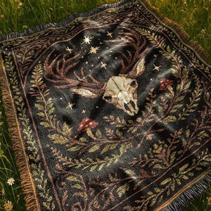 Könnte beinhalten: Schwarze gewebte Decke mit einem Hirschschädel, Sternen und botanischen Elementen. Das Design zeigt einen Hirschschädel mit Geweih, Pilzen und Blättern in Braun-, Grün- und Goldtönen. Die Decke hat einen Fransenrand und liegt auf Gras.