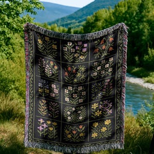 May include: Black woven throw blanket with a floral pattern. The blanket features a grid of squares, each containing a different floral design in shades of yellow, pink, orange, and purple. The edges have a fringed trim.