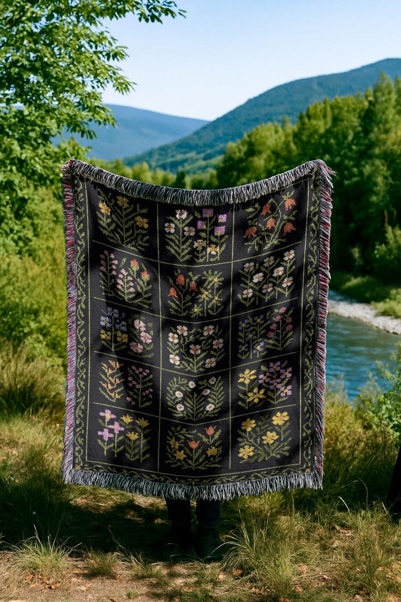 May include: Black woven throw blanket with a floral pattern. The blanket features a grid of squares, each containing a different floral design in shades of yellow, pink, orange, and purple. The edges have a fringed trim.