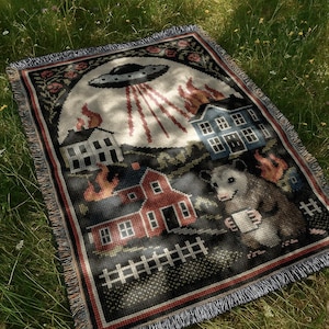 May include: Woven throw blanket with a whimsical design featuring a UFO, burning houses, and an opossum holding a mug. The blanket has a black border with red and white accents and fringe.