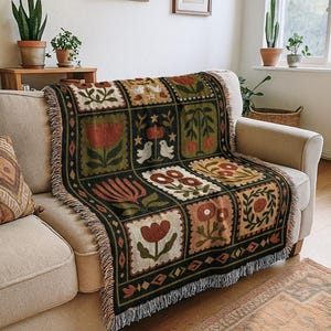 May include: A woven throw blanket with a floral pattern in shades of red, green, and beige is draped over a beige sofa. The blanket features a repeating square design with various flower and bird motifs. The edges are finished with a fringe.