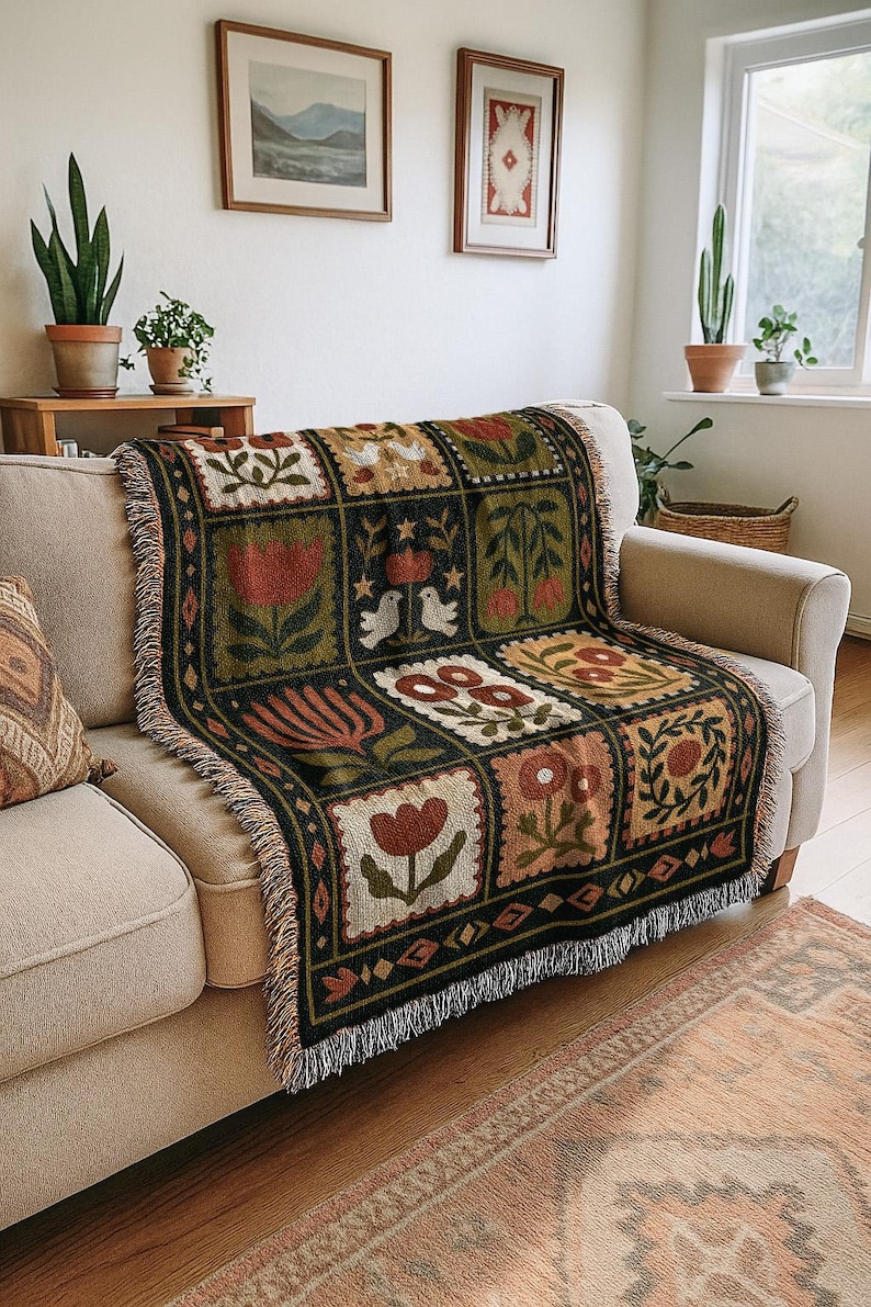 May include: A woven throw blanket with a floral pattern in shades of red, green, and beige is draped over a beige sofa. The blanket features a repeating square design with various flower and bird motifs. The edges are finished with a fringe.