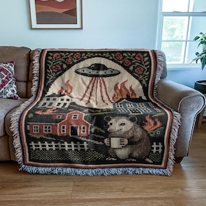 May include: Woven throw blanket featuring a whimsical design with a UFO, burning buildings, and an opossum holding a mug. The blanket has a black border with a fringe and is draped over a brown sofa.
