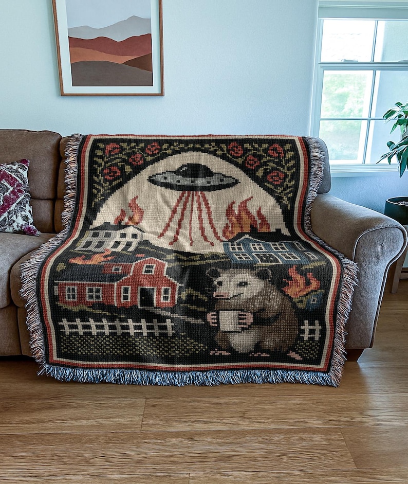 May include: Woven throw blanket featuring a whimsical design with a UFO, burning buildings, and an opossum holding a mug. The blanket has a black border with a fringe and is draped over a brown sofa.
