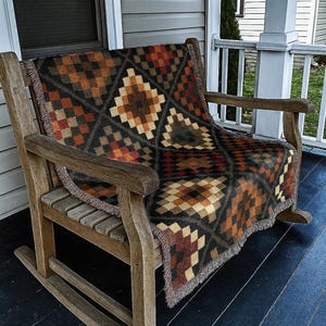 May include: A wooden rocking chair with a patterned blanket draped over it. The blanket features a geometric design in shades of brown, orange, and black. The chair is on a porch with a gray exterior and white trim.