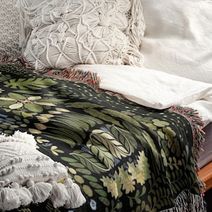 May include: A black throw blanket featuring a botanical design of green leaves and flowers, with fringed edges, and a white macrame pillow. The blanket is on a bed with white sheets and a brown headboard.