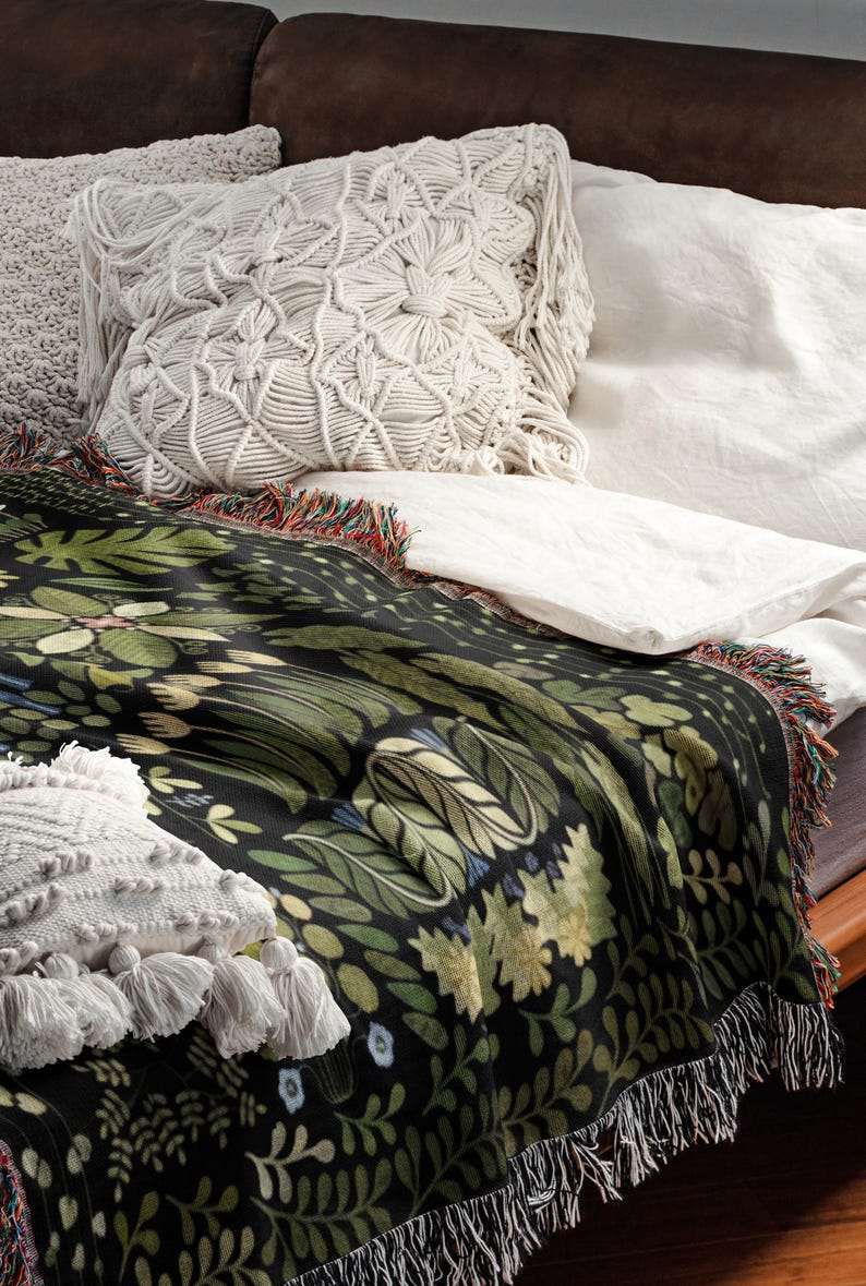 May include: A black throw blanket featuring a botanical design of green leaves and flowers, with fringed edges, and a white macrame pillow. The blanket is on a bed with white sheets and a brown headboard.